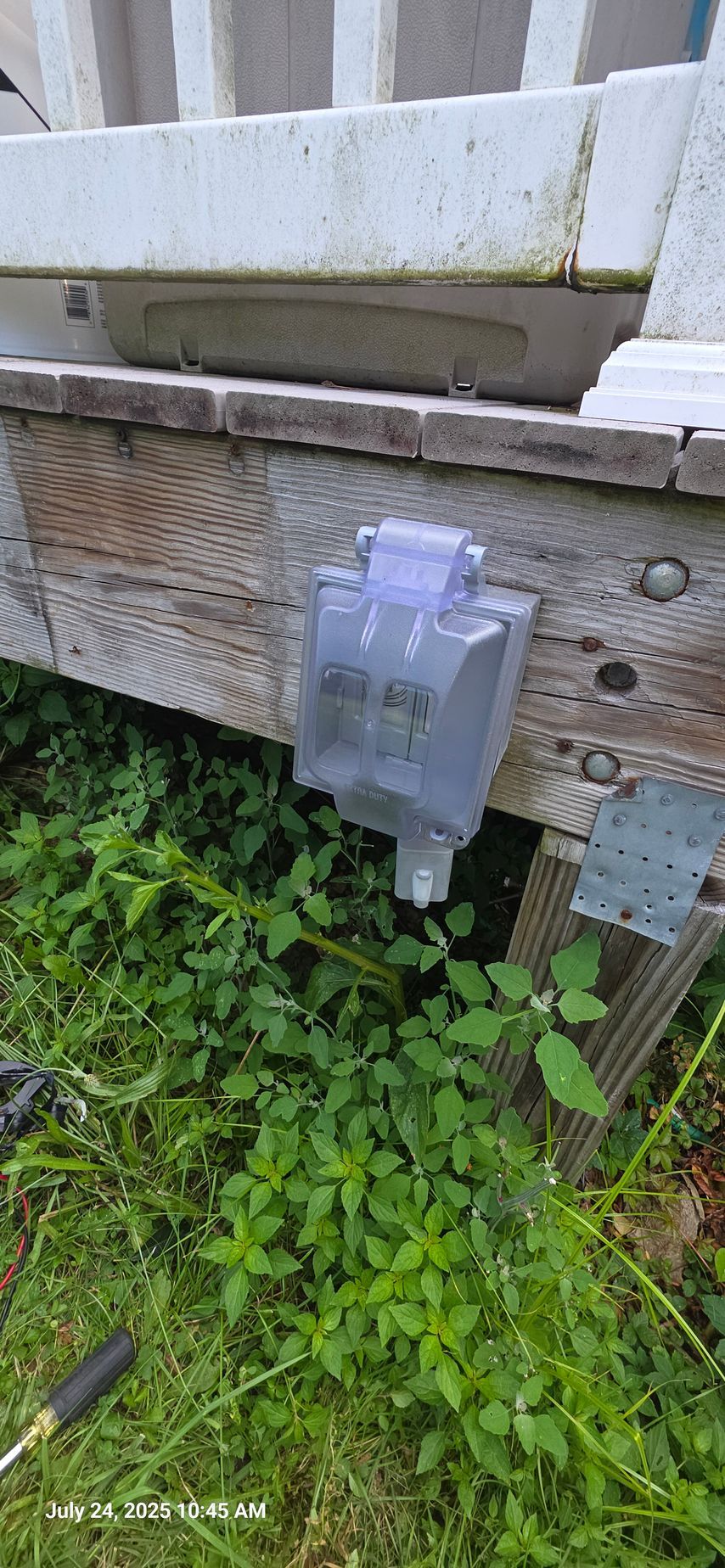 An outdoor electrical outlet is mounted on weathered wooden siding, protected by a clear plastic cover. Green grass surrounds it.