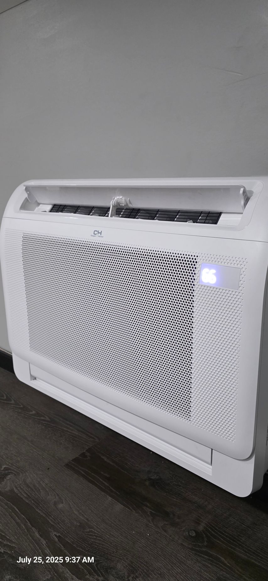 White, window air conditioner with digital display. Leaning against a gray wall. Dark floor.