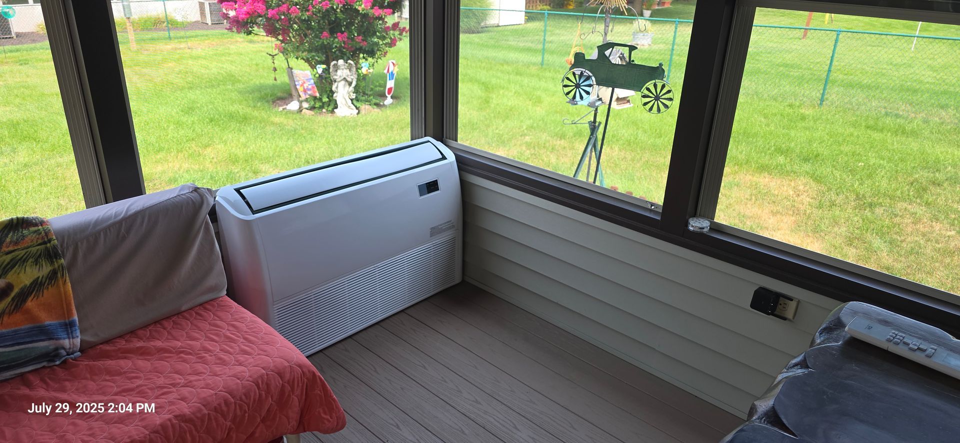 Sunroom with air conditioner, cushions, and view of a green yard with a decorative wagon.