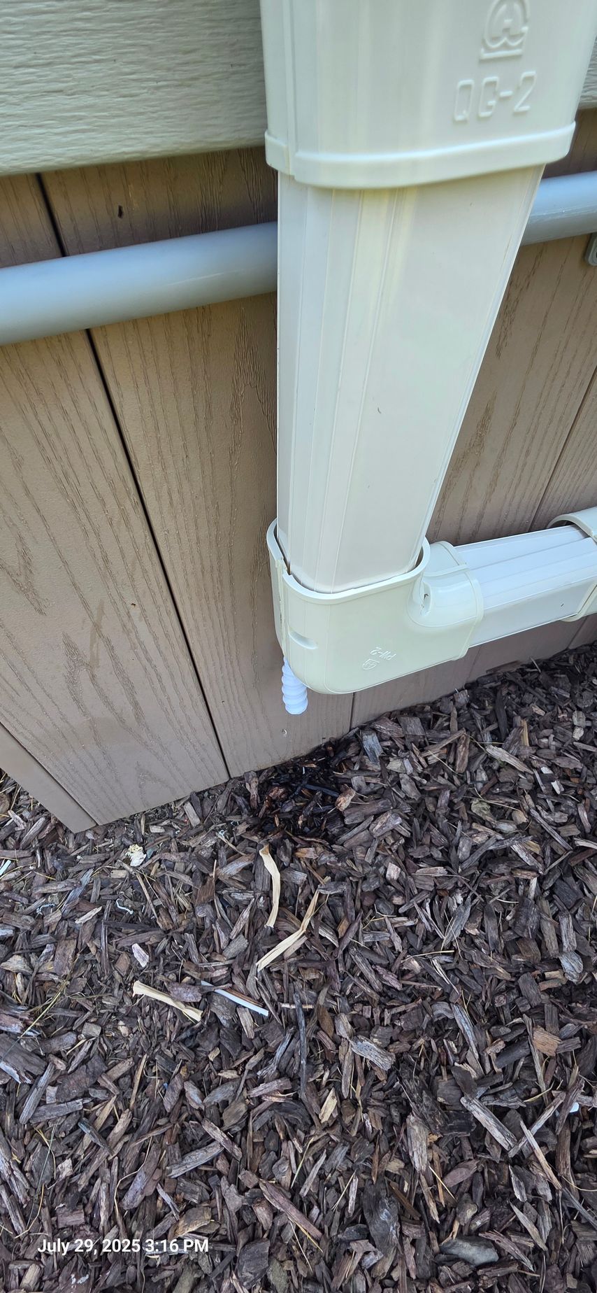 A downspout, connected to a gutter, near a wood-sided building, empties into mulch.