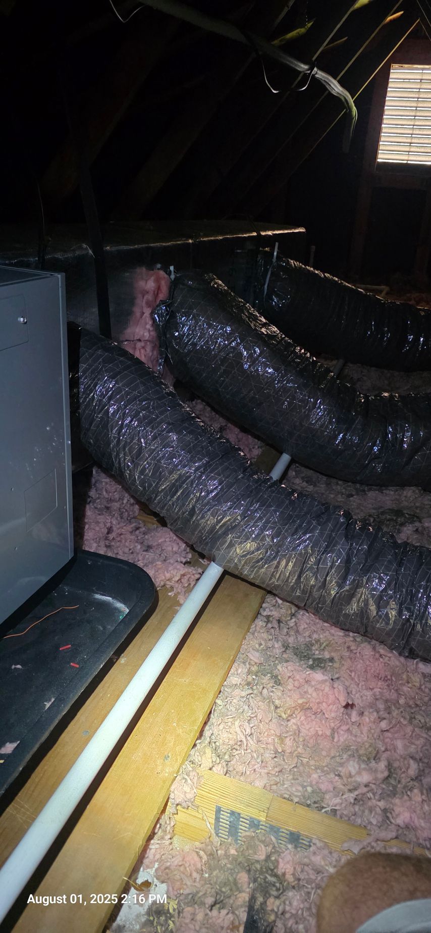 An attic with black flexible ductwork, pink insulation, and a white pipe.