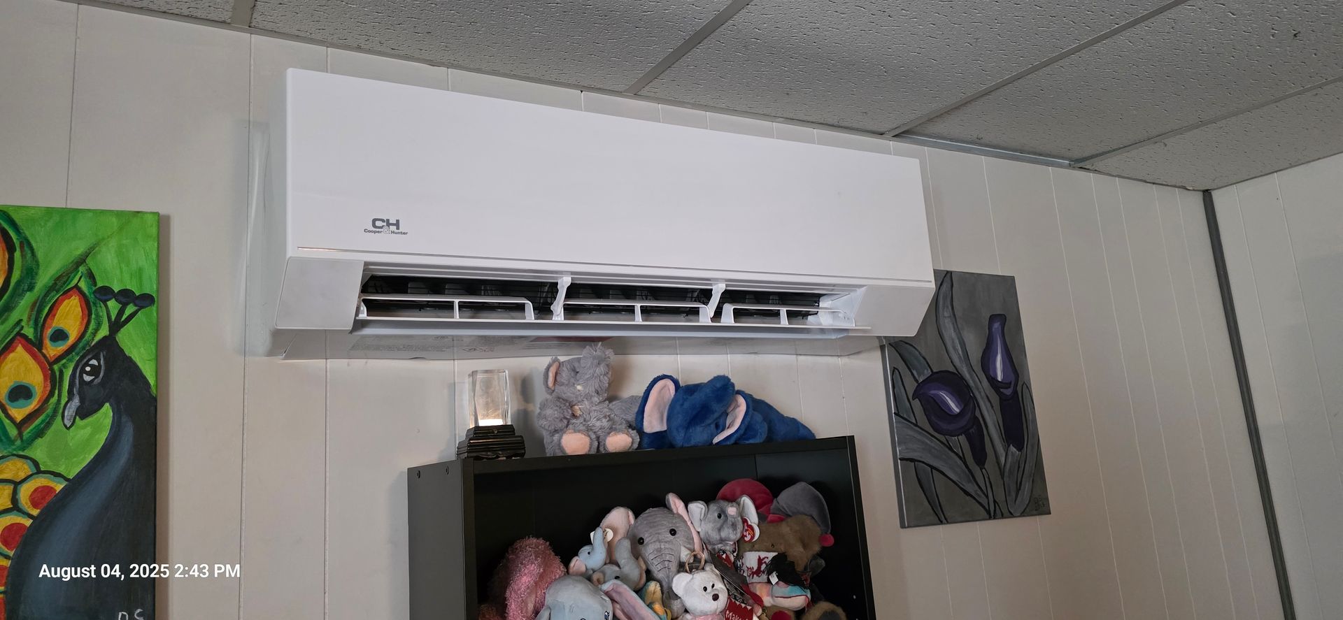 White air conditioner on a wall above a shelf with stuffed animals; artwork on the wall.