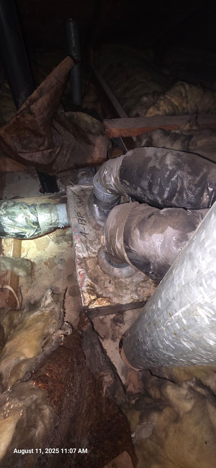 Ductwork and insulation in an attic, with various pipes and vents visible. Dark and dusty environment.
