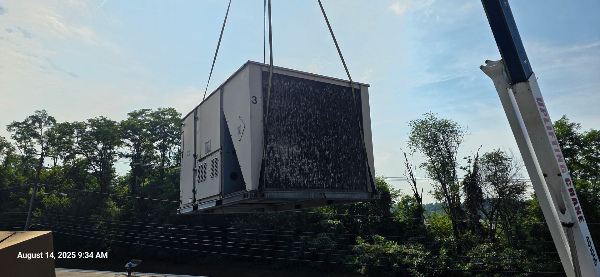 A large HVAC unit is lifted by a crane over trees on a sunny day.