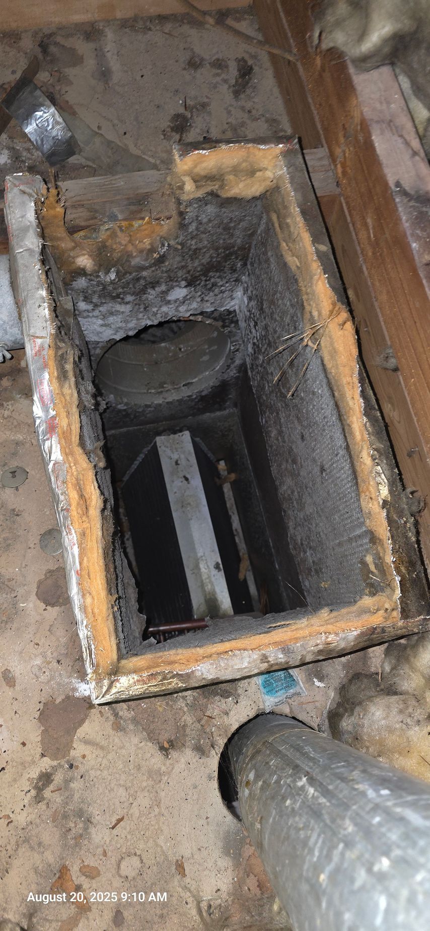 An open, rectangular duct in a dusty attic, with a round ventilation opening. A metallic duct is on the floor.