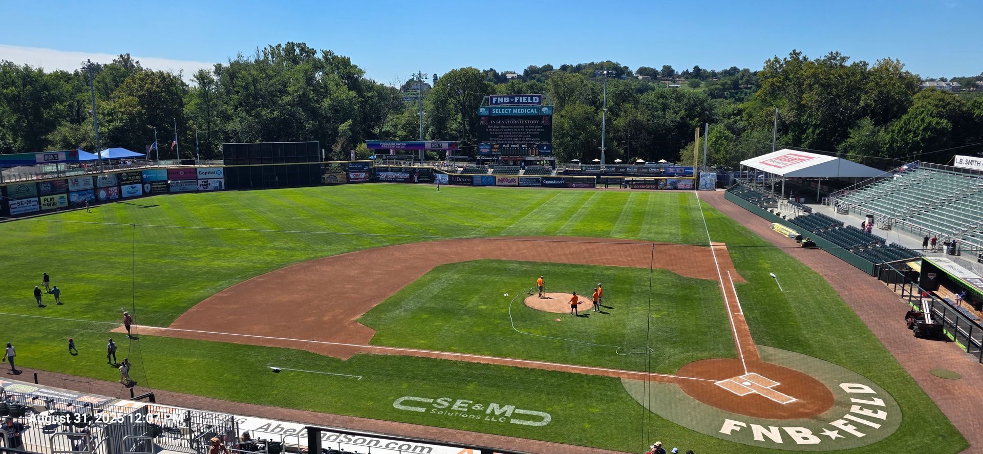 Baseball field with green grass, brown dirt, and a scoreboard, under a blue sky.