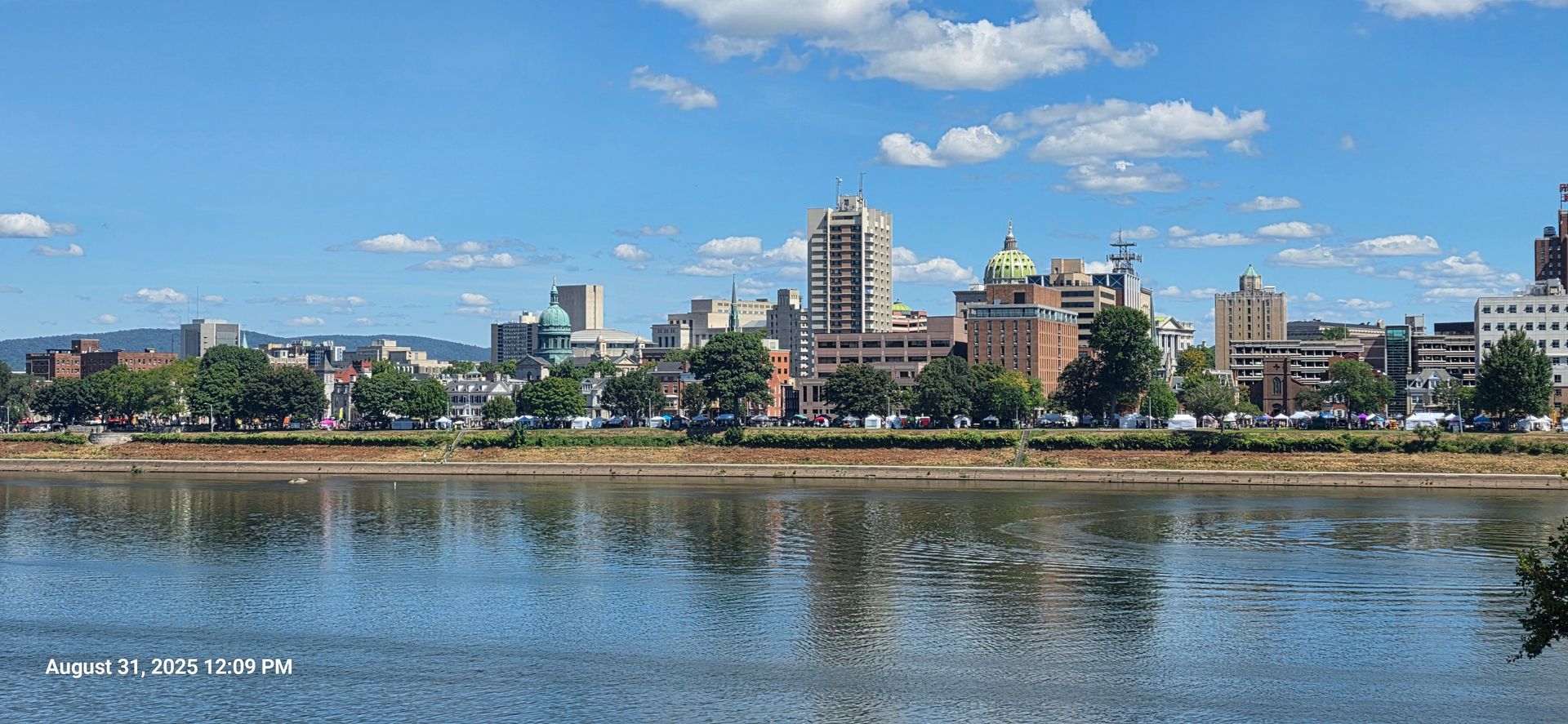 City skyline reflected in water, trees along the shore, under a blue sky with white clouds.