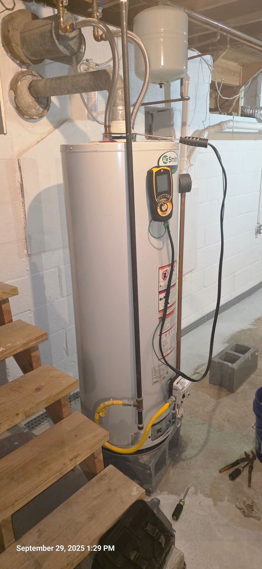 A gray water heater in a basement. Yellow gas line. Stairs and cinder blocks are nearby. A digital gauge is attached.