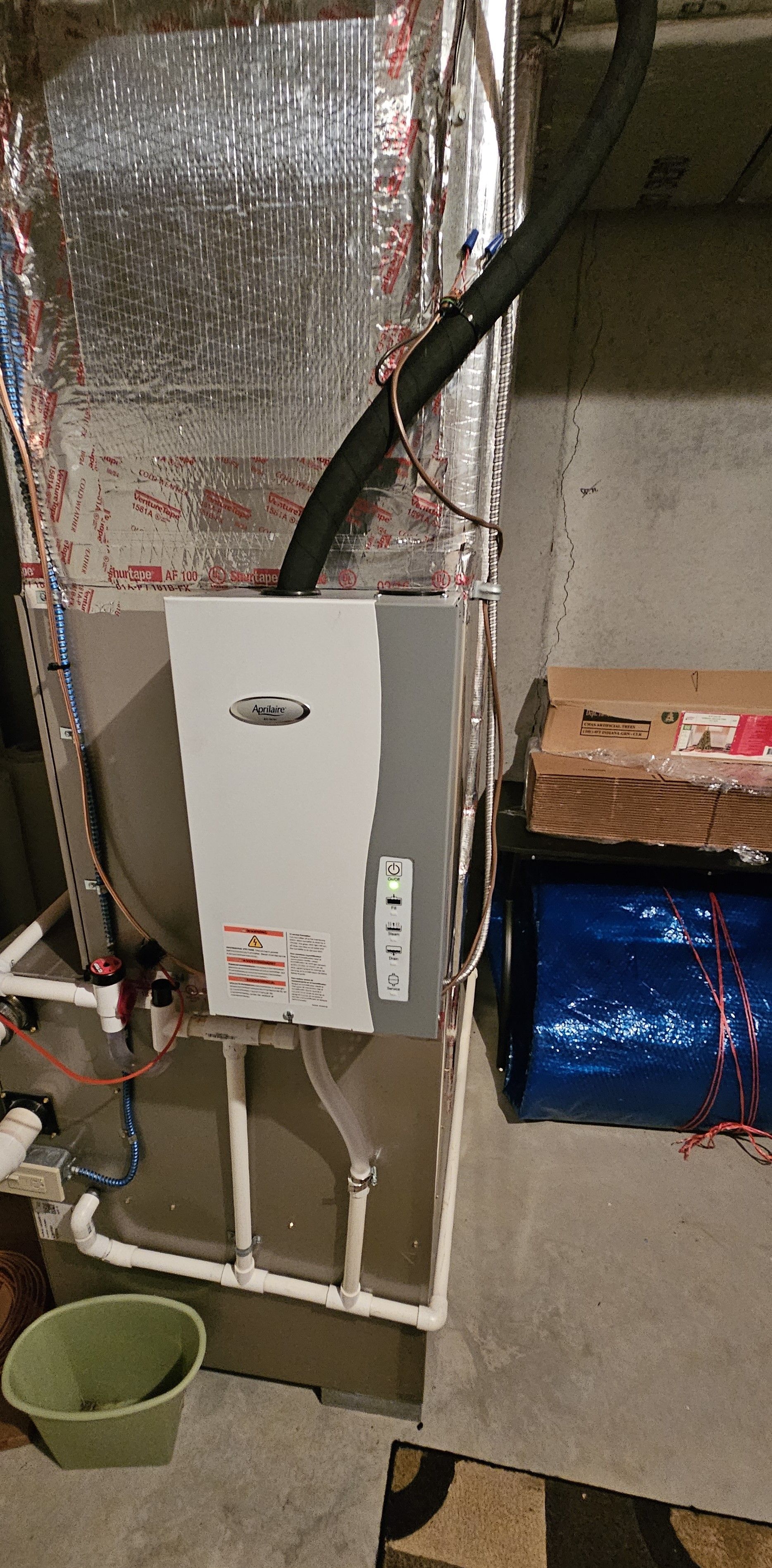 A water heater and ductwork in a basement. A black hose runs from the top to a box on the floor.