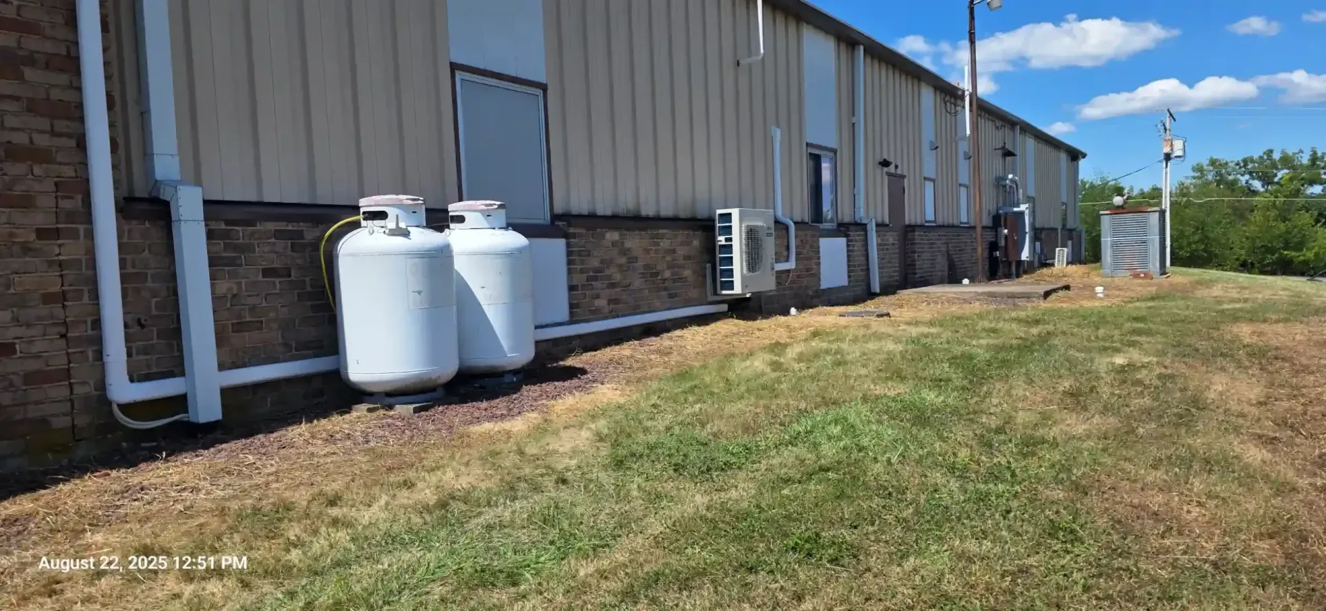 Exterior view of a commercial building with propane tanks, air conditioning units, and a grassy area. Blue sky.