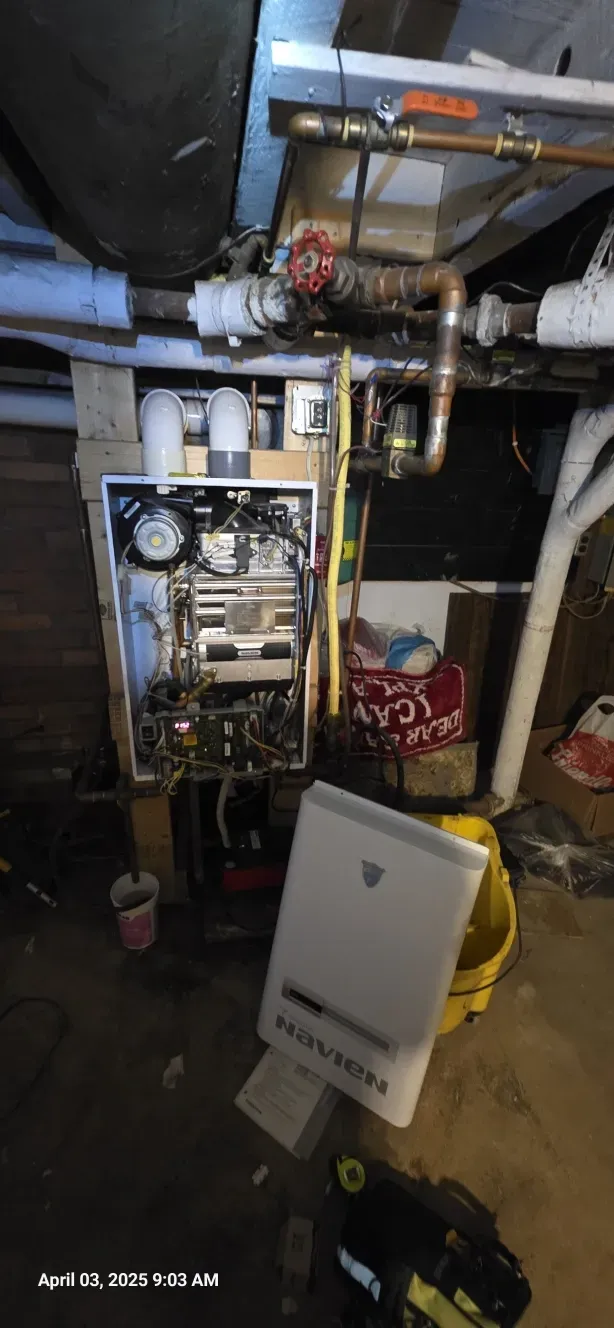 A wall-mounted heating system with exposed components, plumbing, and a white control panel in a basement.