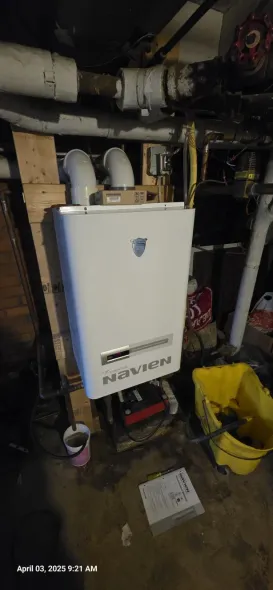 A white wall-mounted boiler in a basement. Pipes above, a yellow bucket, and tools in view.