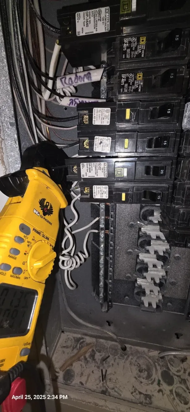 A yellow clamp meter is reading an electrical panel. Wires and circuit breakers are visible.