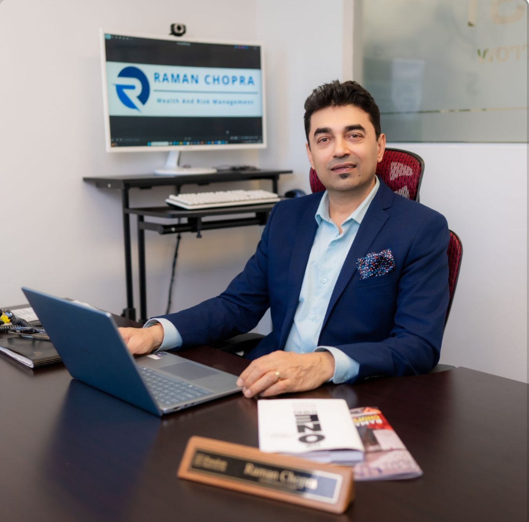 Business professional seated at a desk with laptop in an office, smiling beside a monitor displaying a company logo