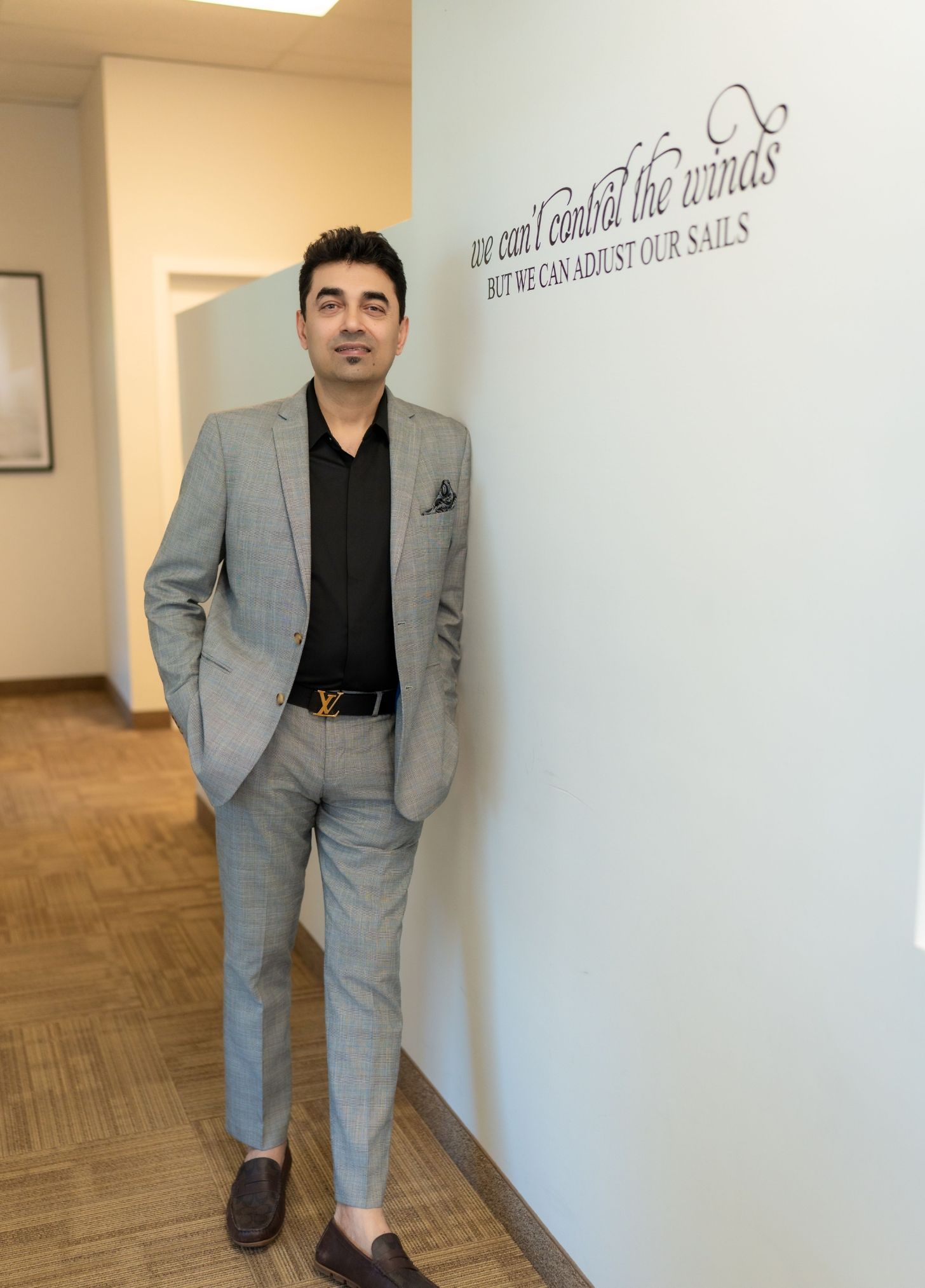 Man in a gray suit leaning against a wall in a hallway beside a clinic sign