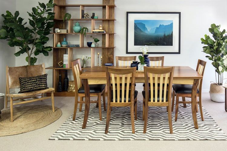 A wooden dining set with chairs on a geometric rug, centered in a room with a large bookshelf and framed landscape art.