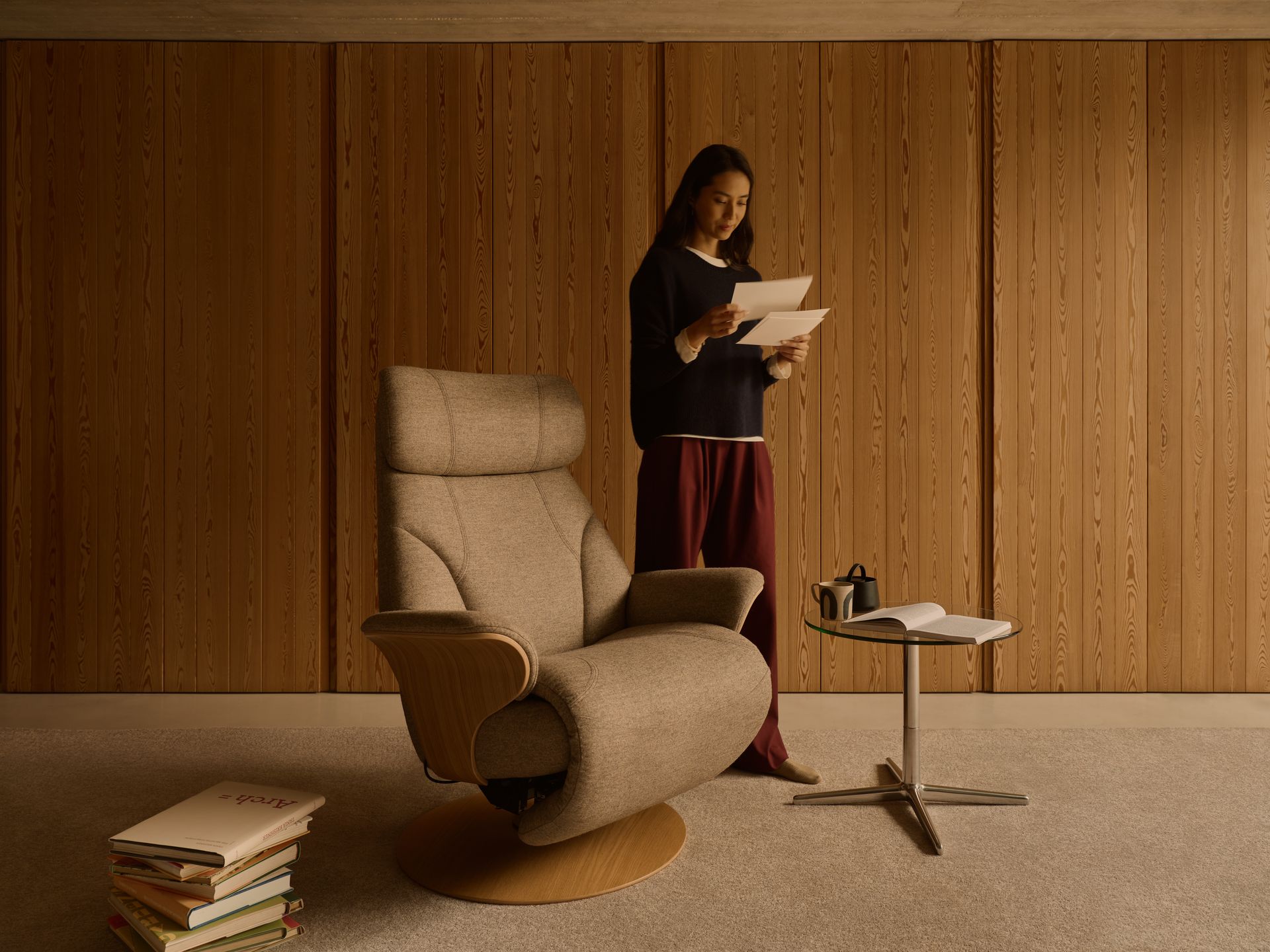 A person in dark clothes stands reading in a room with wood-paneled walls, an armchair, a side table, and stacked books.
