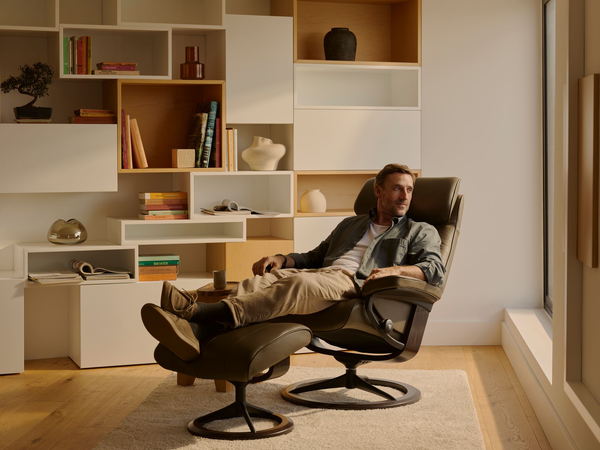 A person relaxes in a dark recliner with a matching footstool in a bright room with contemporary white shelving units.