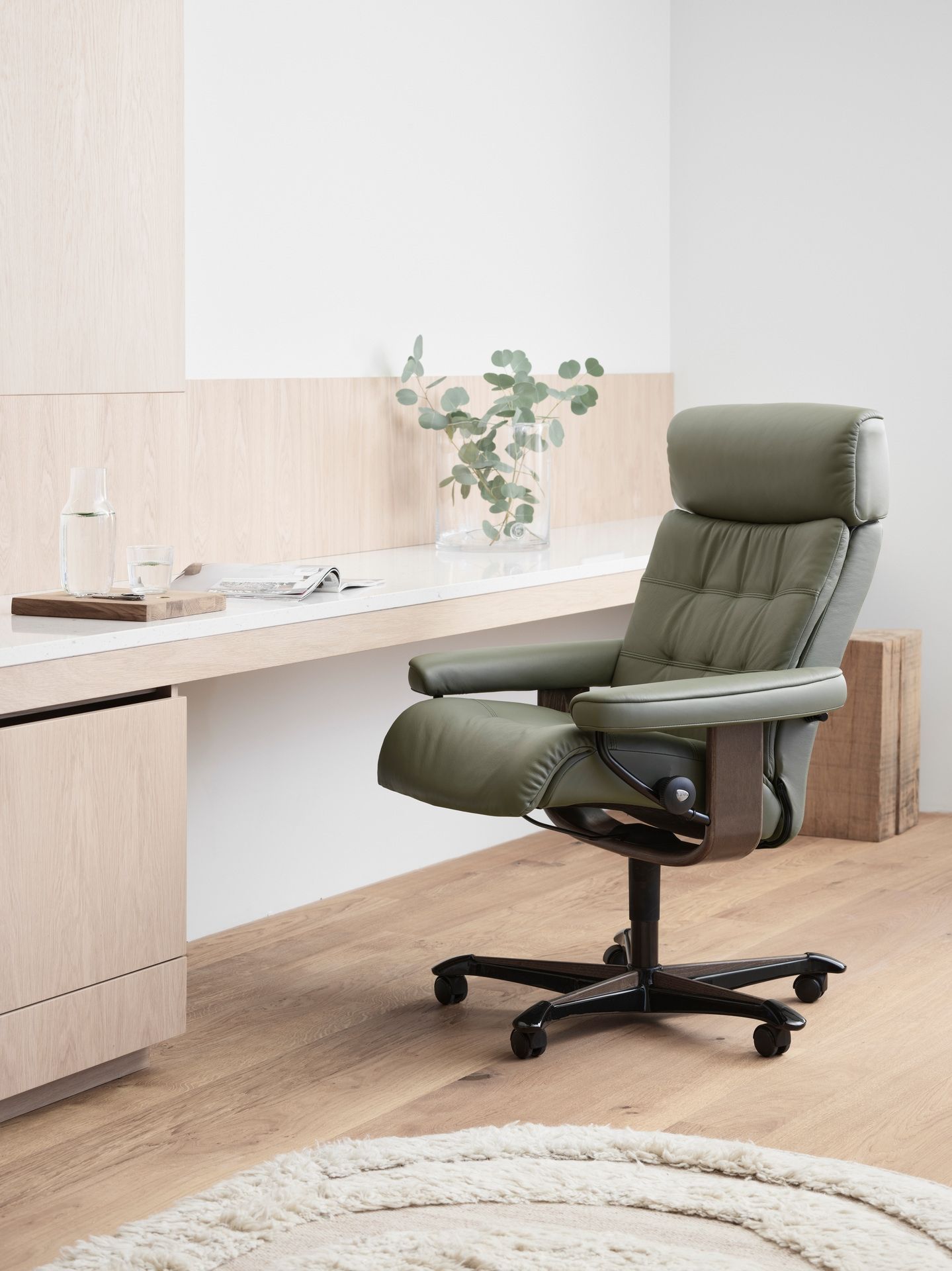 A sage green leather office chair with a reclined footrest sits in a minimalist room with light wood floors and desk.