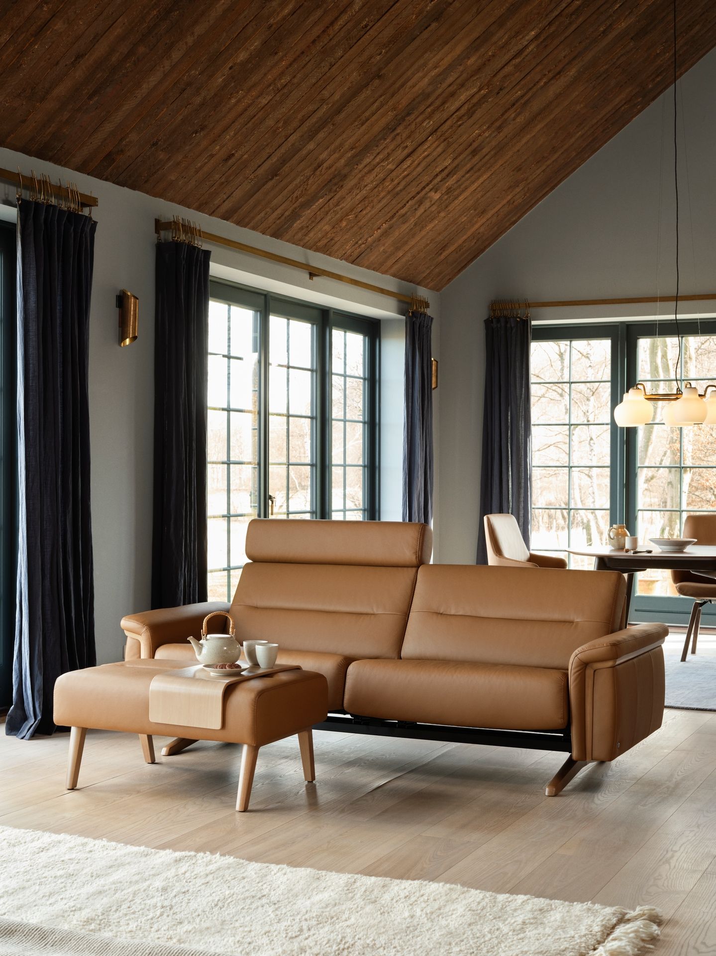A tan leather sofa with a matching ottoman sits in a sunlit room with wooden vaulted ceilings and dark curtains.