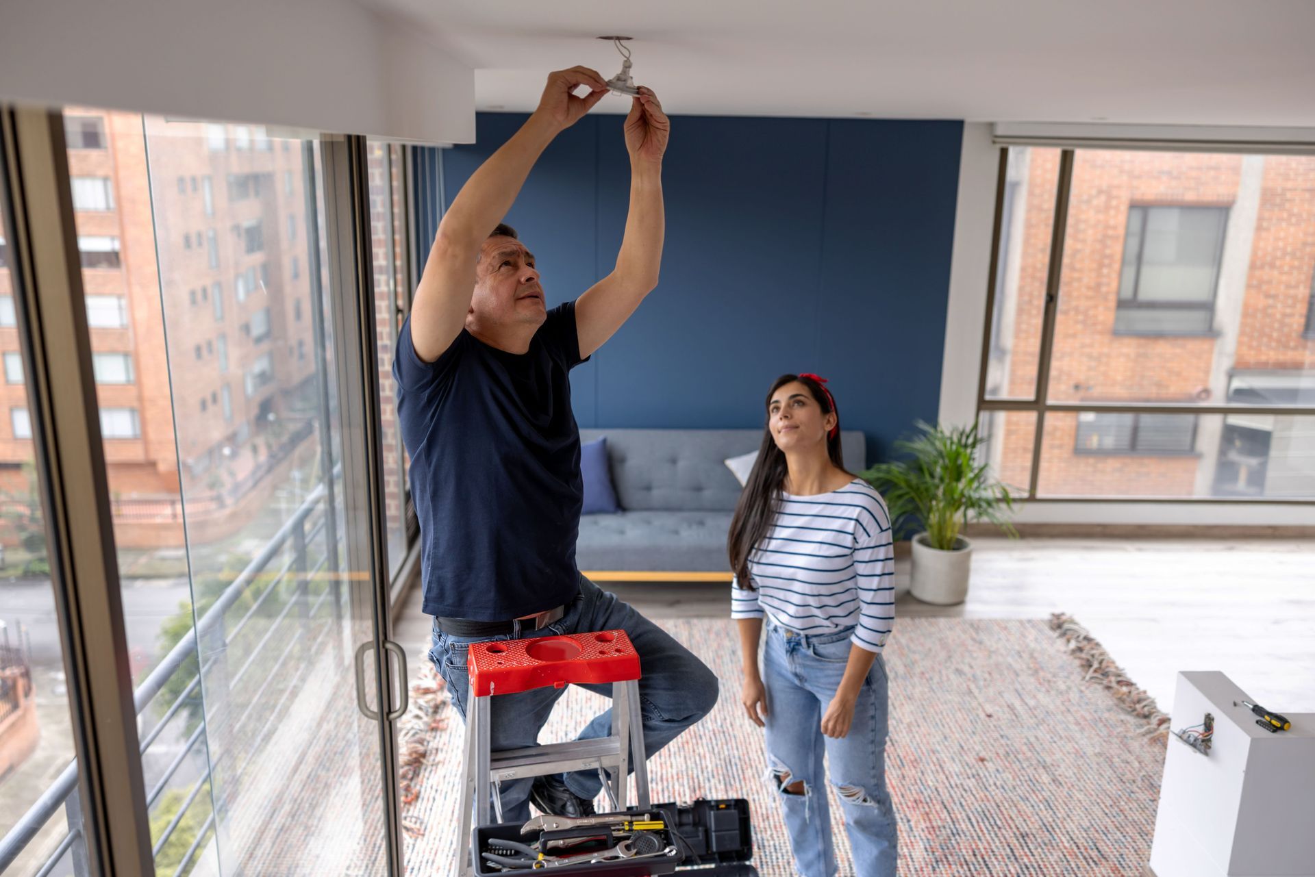 A man is sitting on a ladder fixing a light in a living room while a woman looks on.