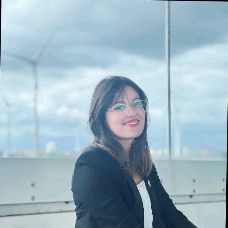 Woman with glasses smiles, wearing a black blazer, in front of a glass window overlooking a cityscape.