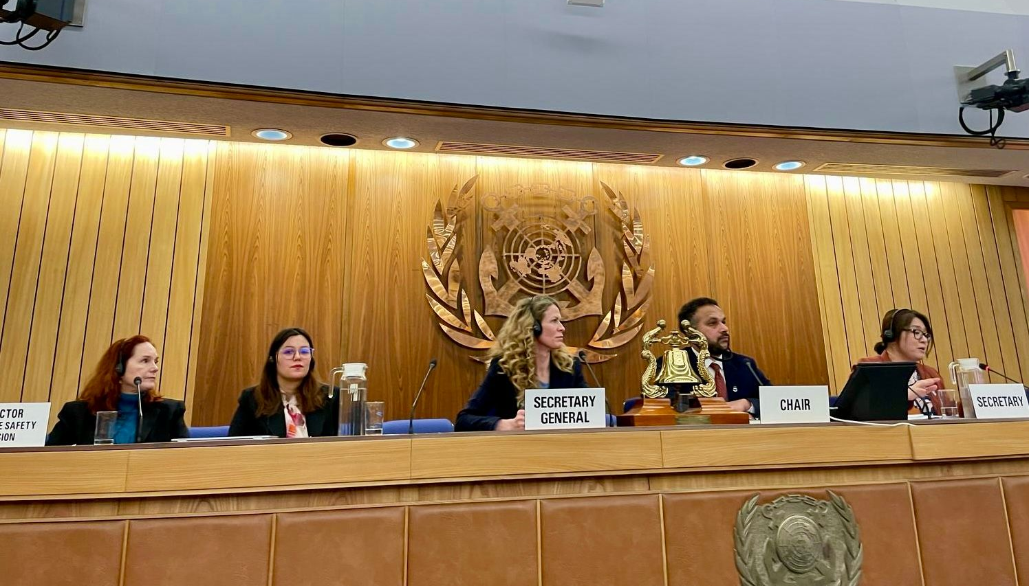 Panel of five people at a wooden desk in a conference room, discussing. A gold seal decorates the wall.