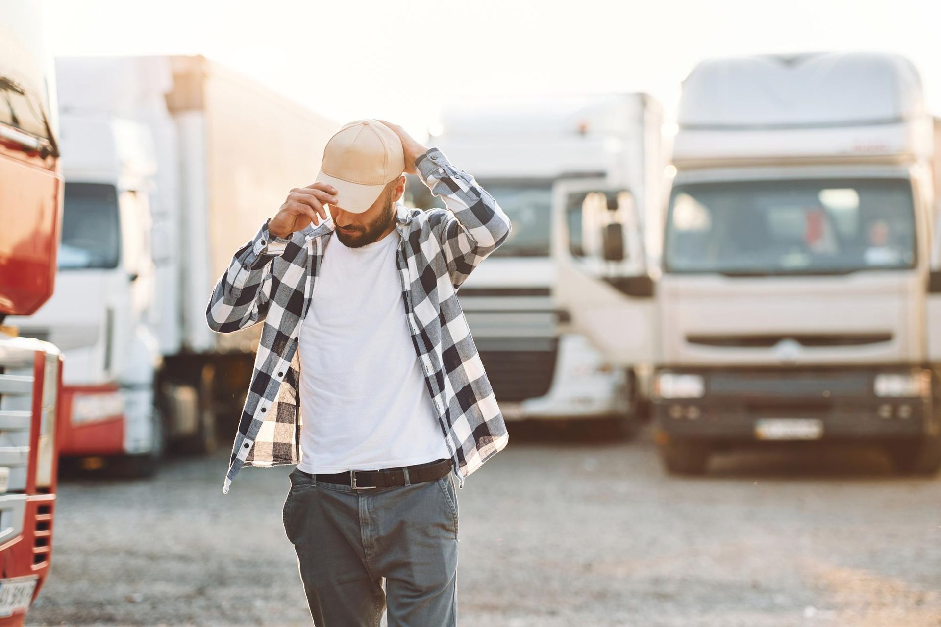 Truck driver adjusting cap in a truck parking lot, trucks in the background. 