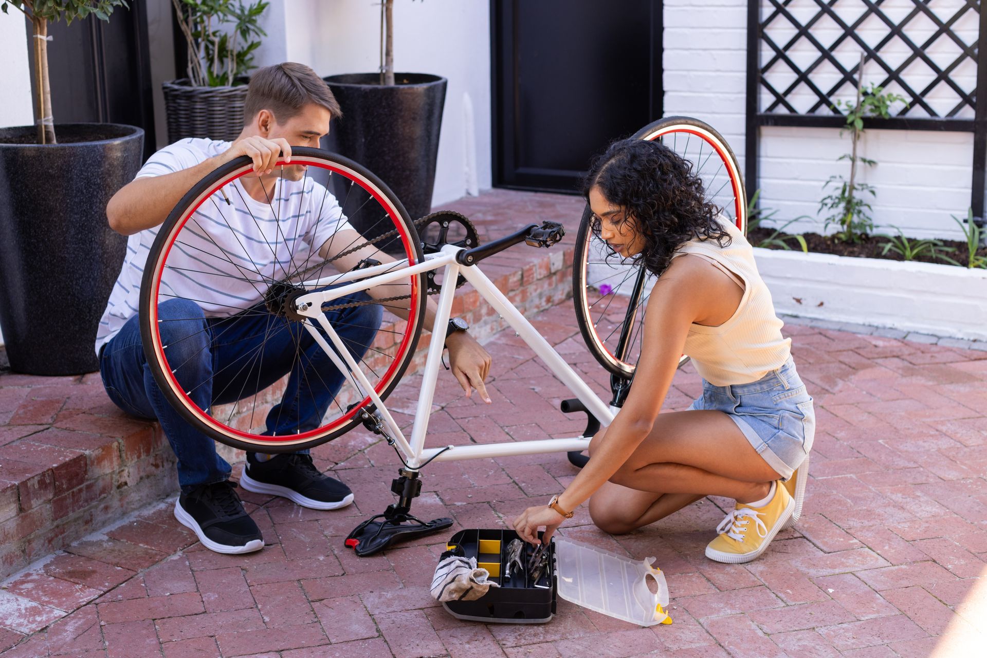 Couple fixing a white bicycle outdoors. One holds a tire, the other examines a toolbox on the brick patio.