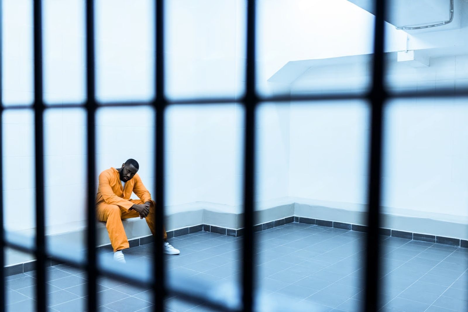 Person in orange jumpsuit sits alone in a cell, behind bars.