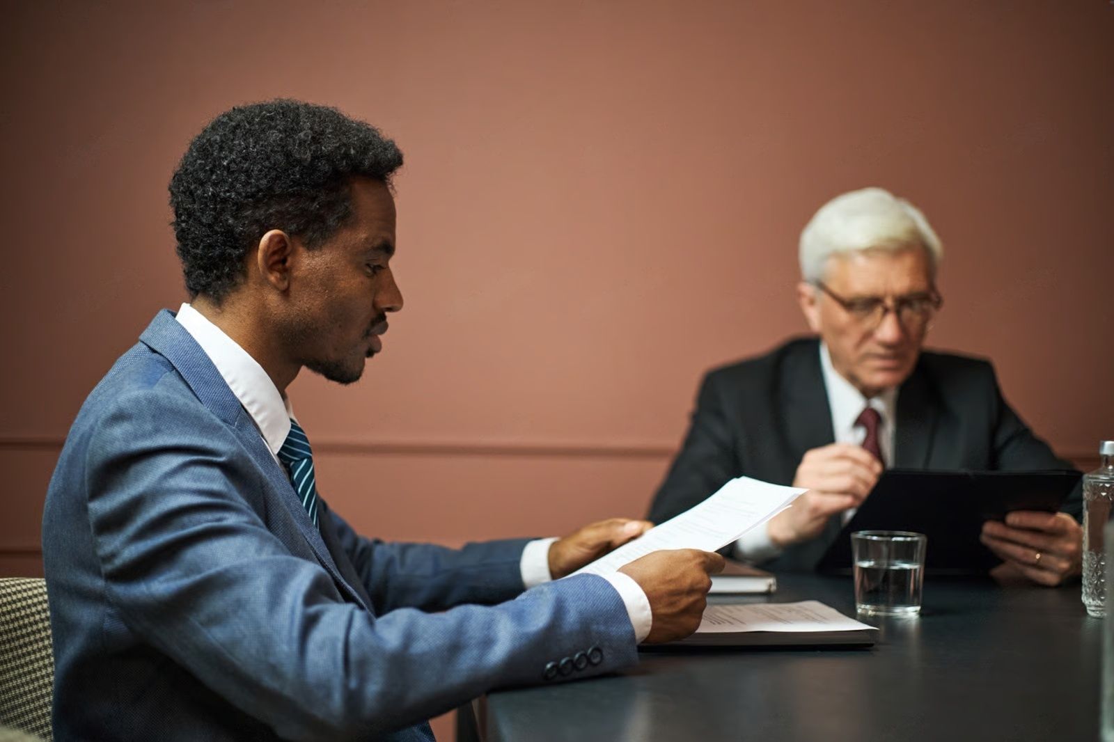 Man in blue suit reviews documents at a table; another man in suit, with glasses, looks at a tablet.