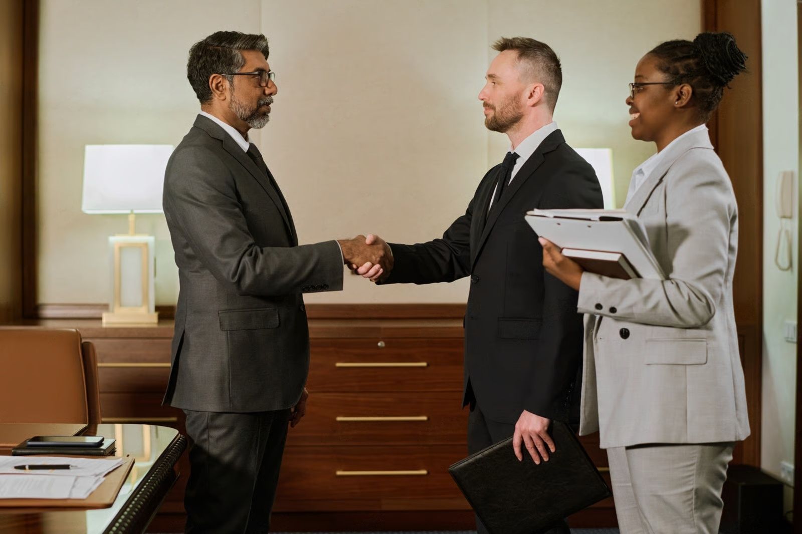 Two men shaking hands in a business setting, with a woman holding a tablet looking on.