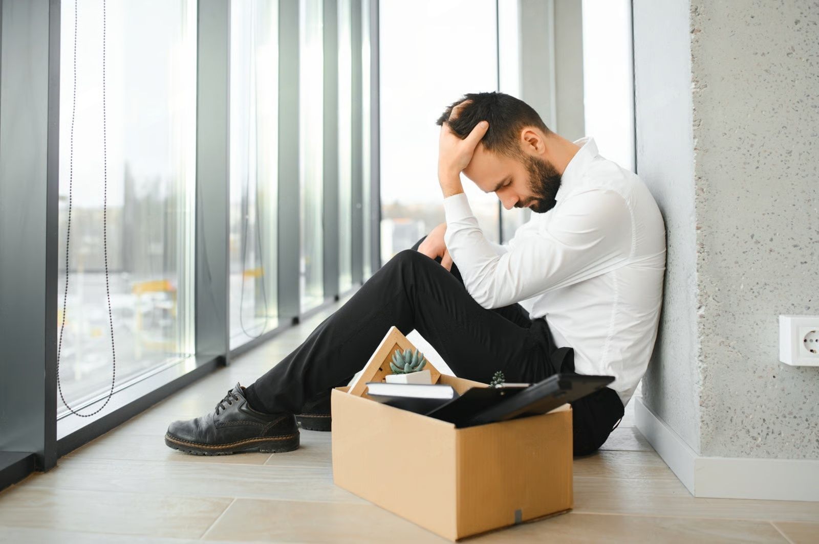 Man sits dejectedly with head in hands next to a box of belongings in an office setting.