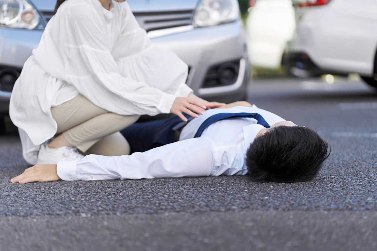 Woman kneeling beside a person lying on asphalt next to a silver car; possible car accident.