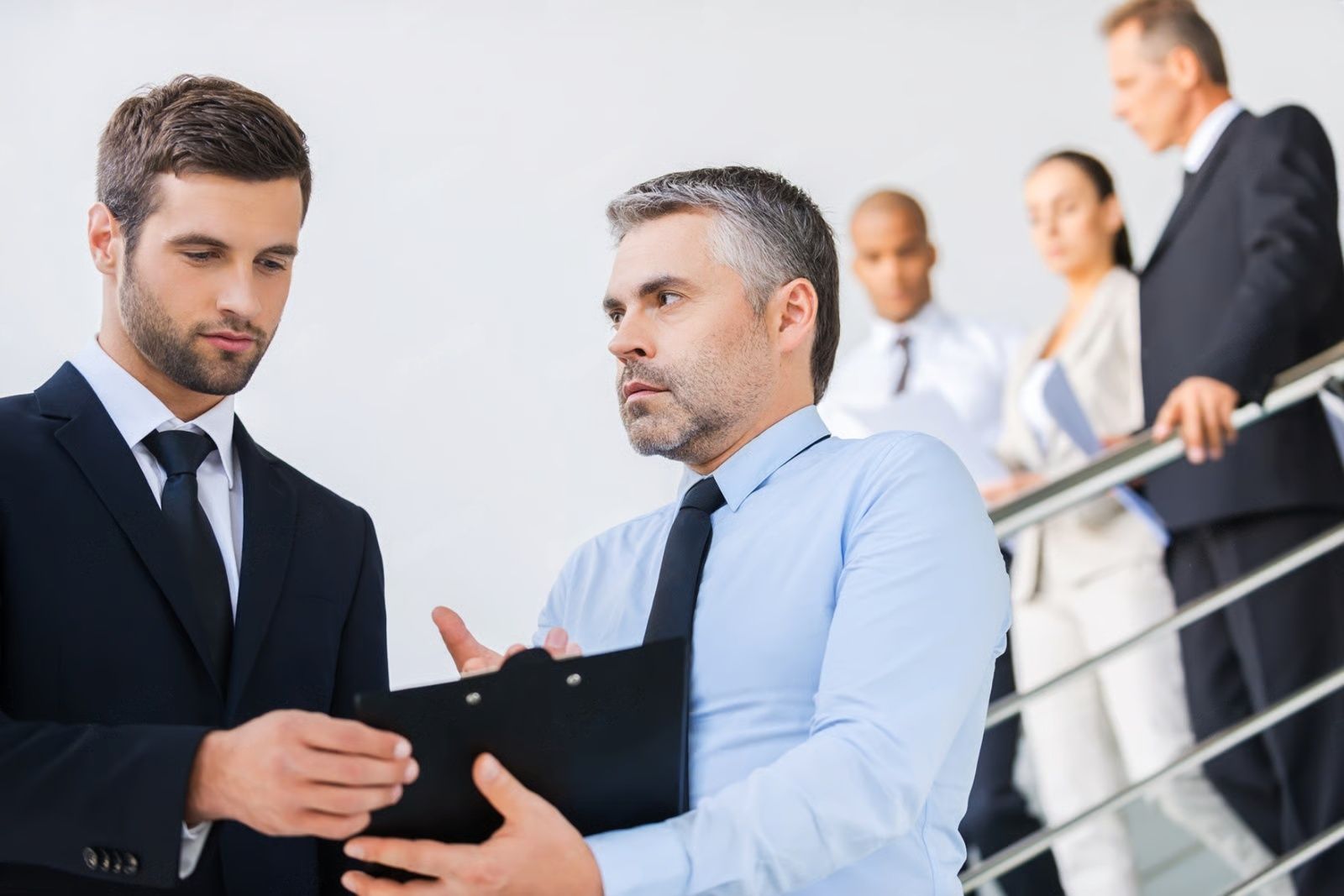 Two men in suits discussing paperwork, other professionals in the background, near a staircase.