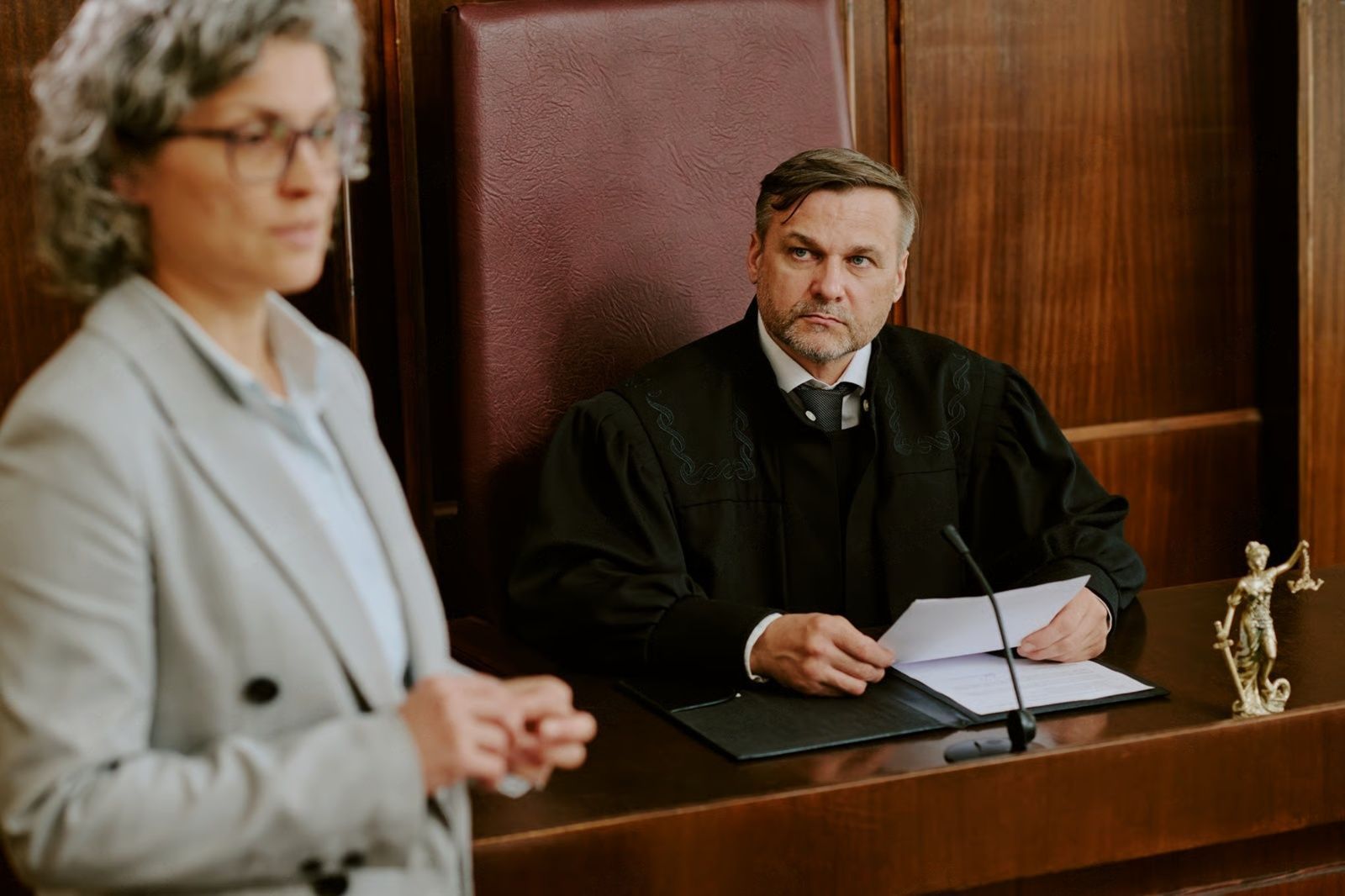 A judge in black robes at a courtroom desk, looking sternly. A lawyer in a gray suit stands before the judge.
