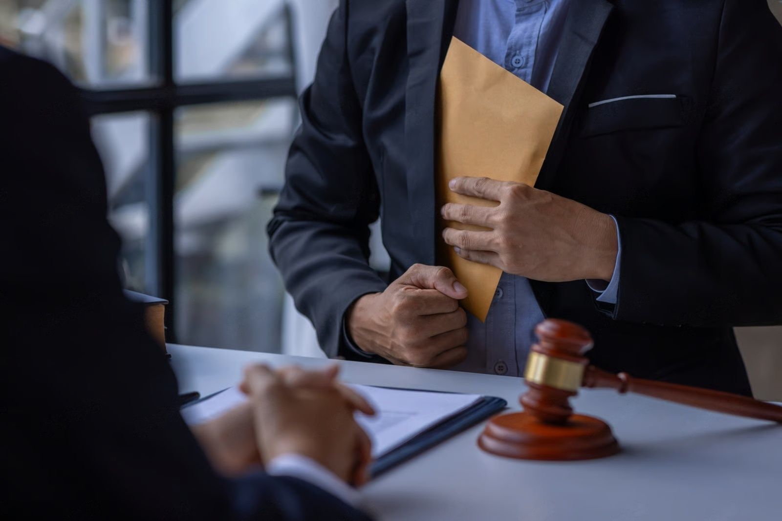 Man in suit hides envelope near a desk with a gavel and document, suggesting bribery.