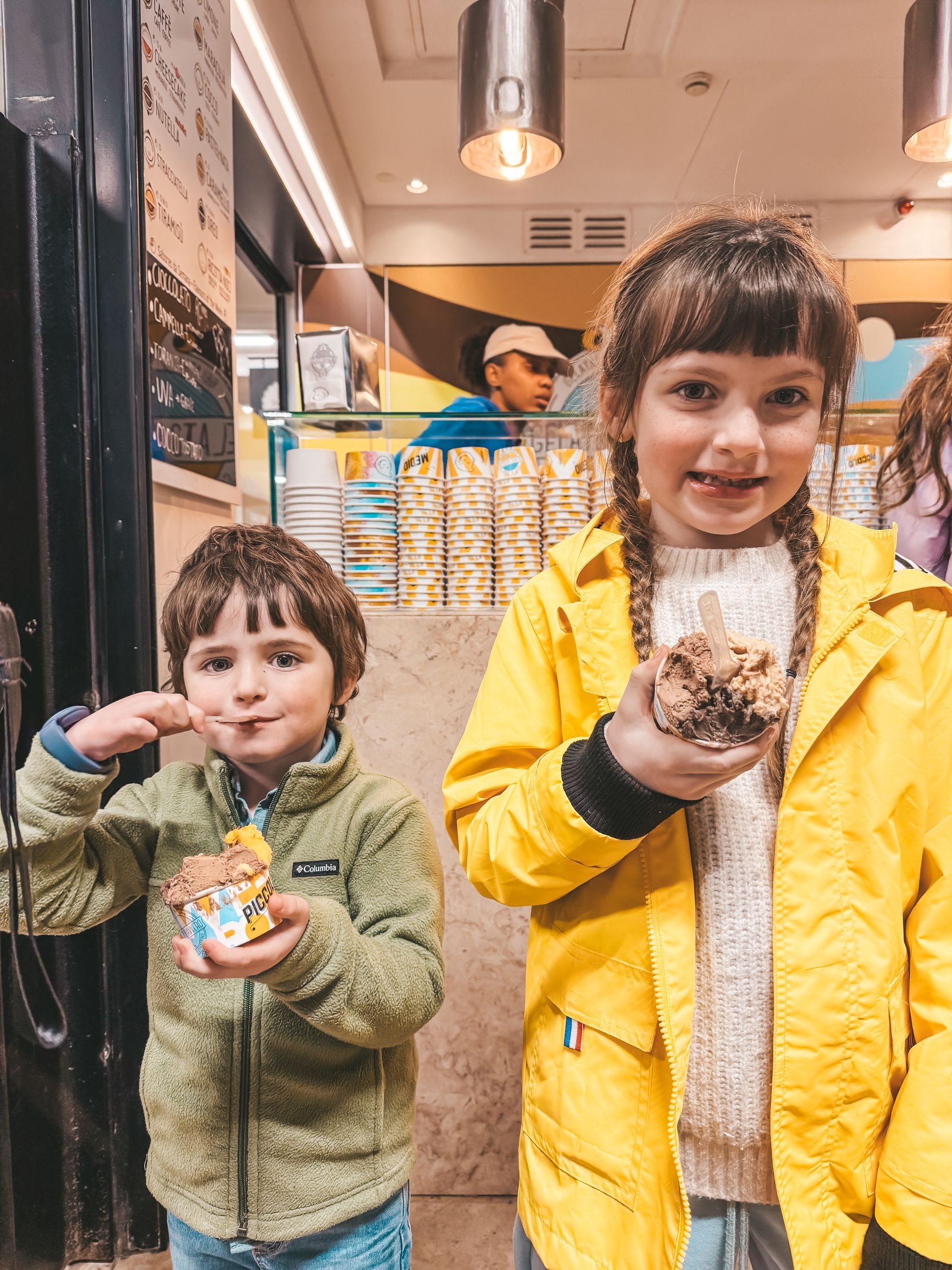 Two children, boy and girl, eating ice cream in a shop. Boy in green jacket, girl in yellow raincoat.