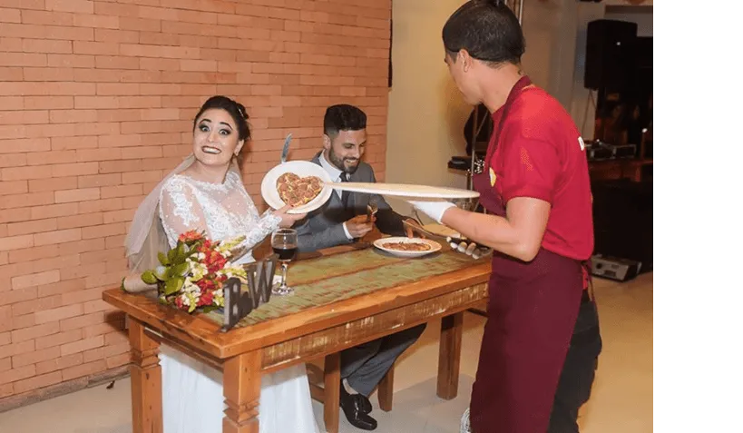 Uma noiva e um noivo estão sentados à mesa enquanto um garçom lhes serve comida.