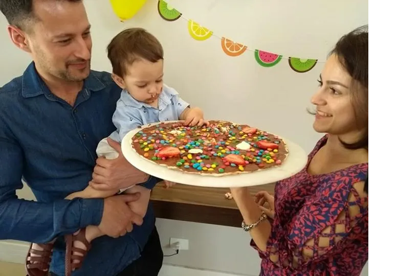 Um homem está segurando um bebê e uma mulher está segurando uma pizza com granulado.