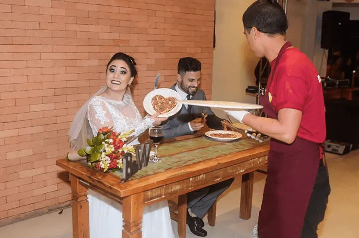Uma noiva e um noivo estão sentados à mesa enquanto um garçom lhes serve comida.