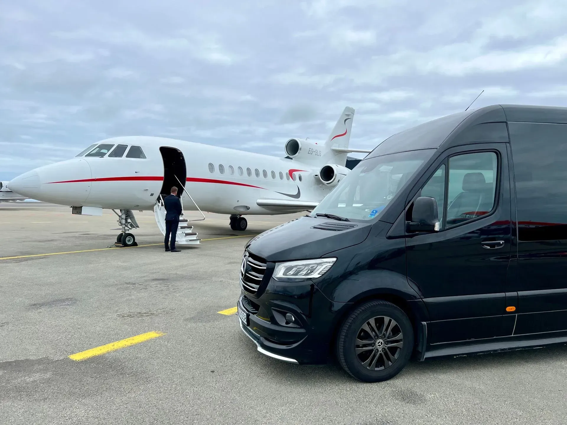Black van parked near a white private jet on an airport tarmac; a person stands near the jet's open door.