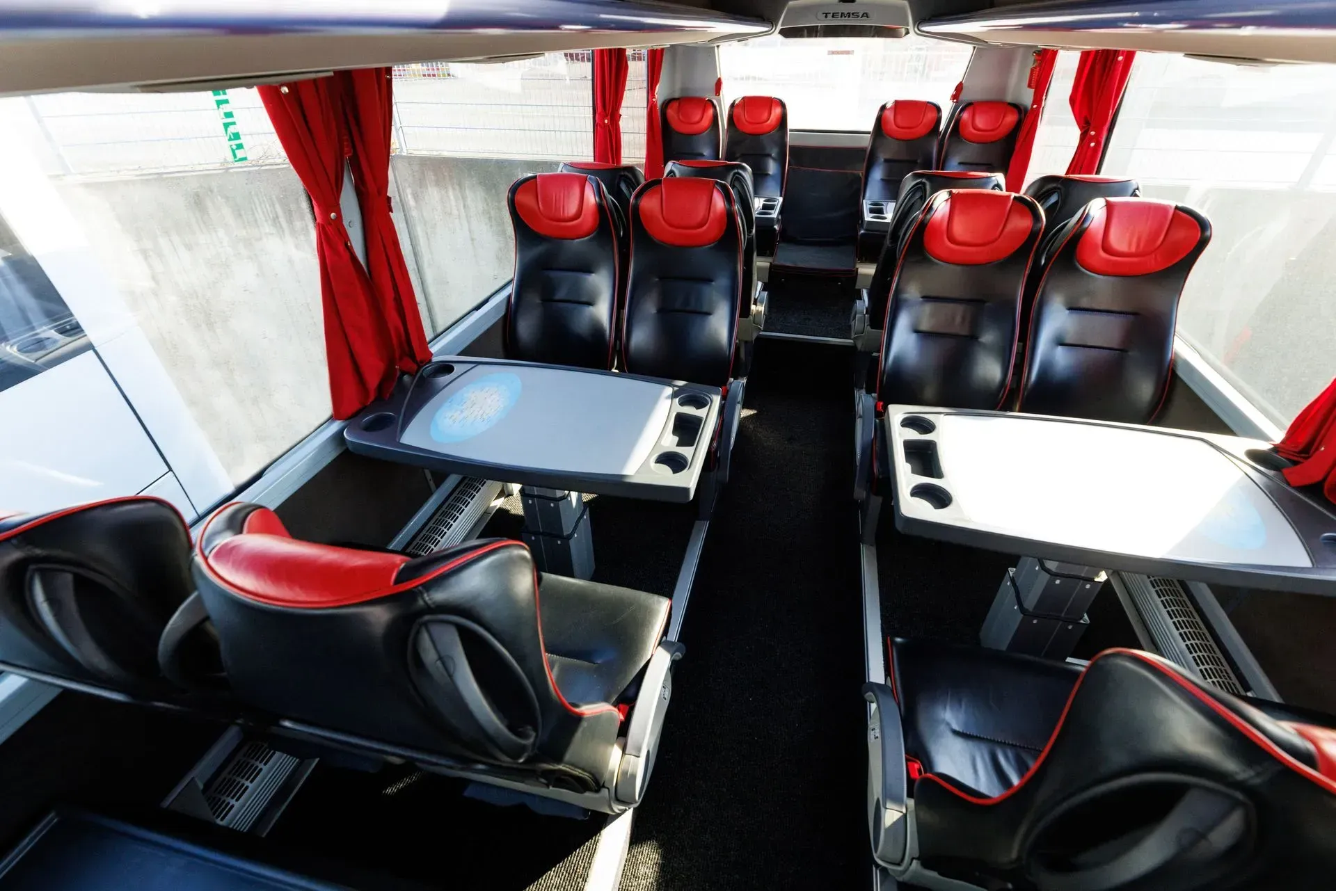 Interior of a bus, rows of black and red seats with tables, red curtains, overhead compartments.