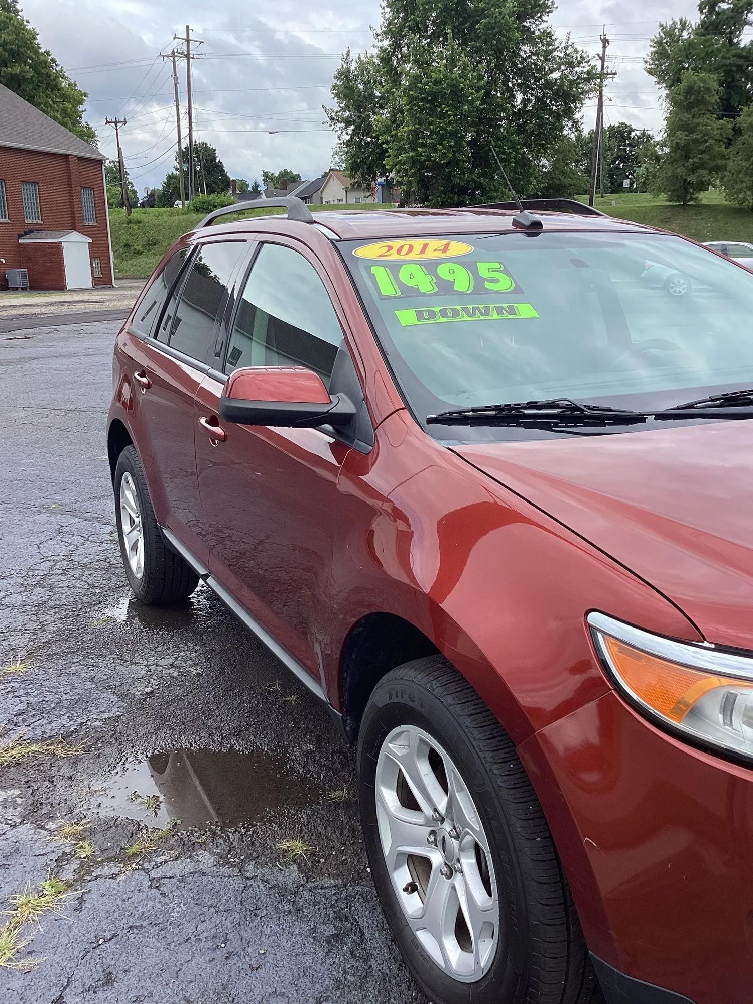 Red SUV with neon green price signs parked on a wet lot near a brick building