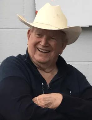 Smiling person in a cream cowboy hat and dark jacket, standing against a light gray wall.