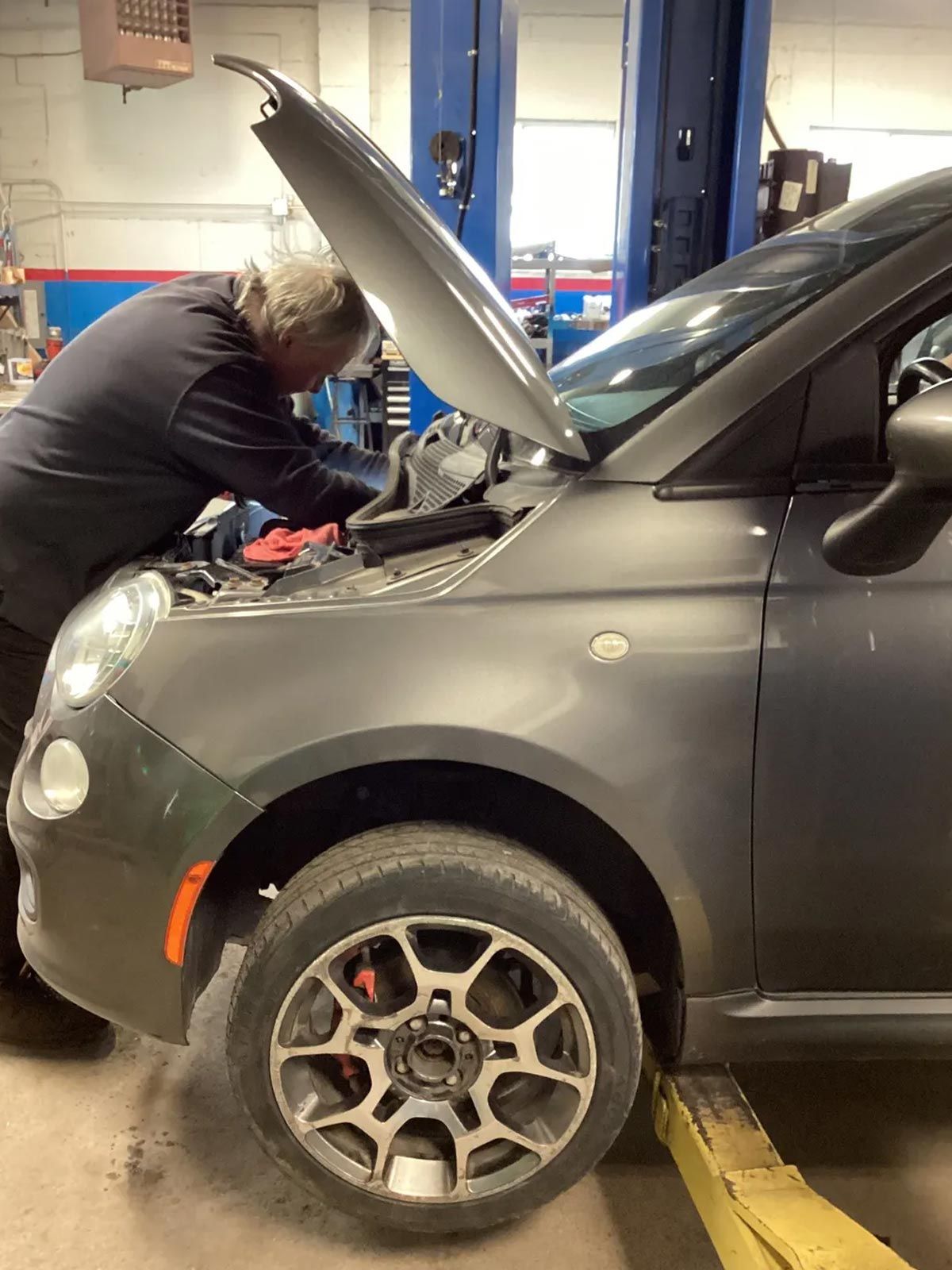 Mechanic working under the open hood of a gray car in an auto repair shop
