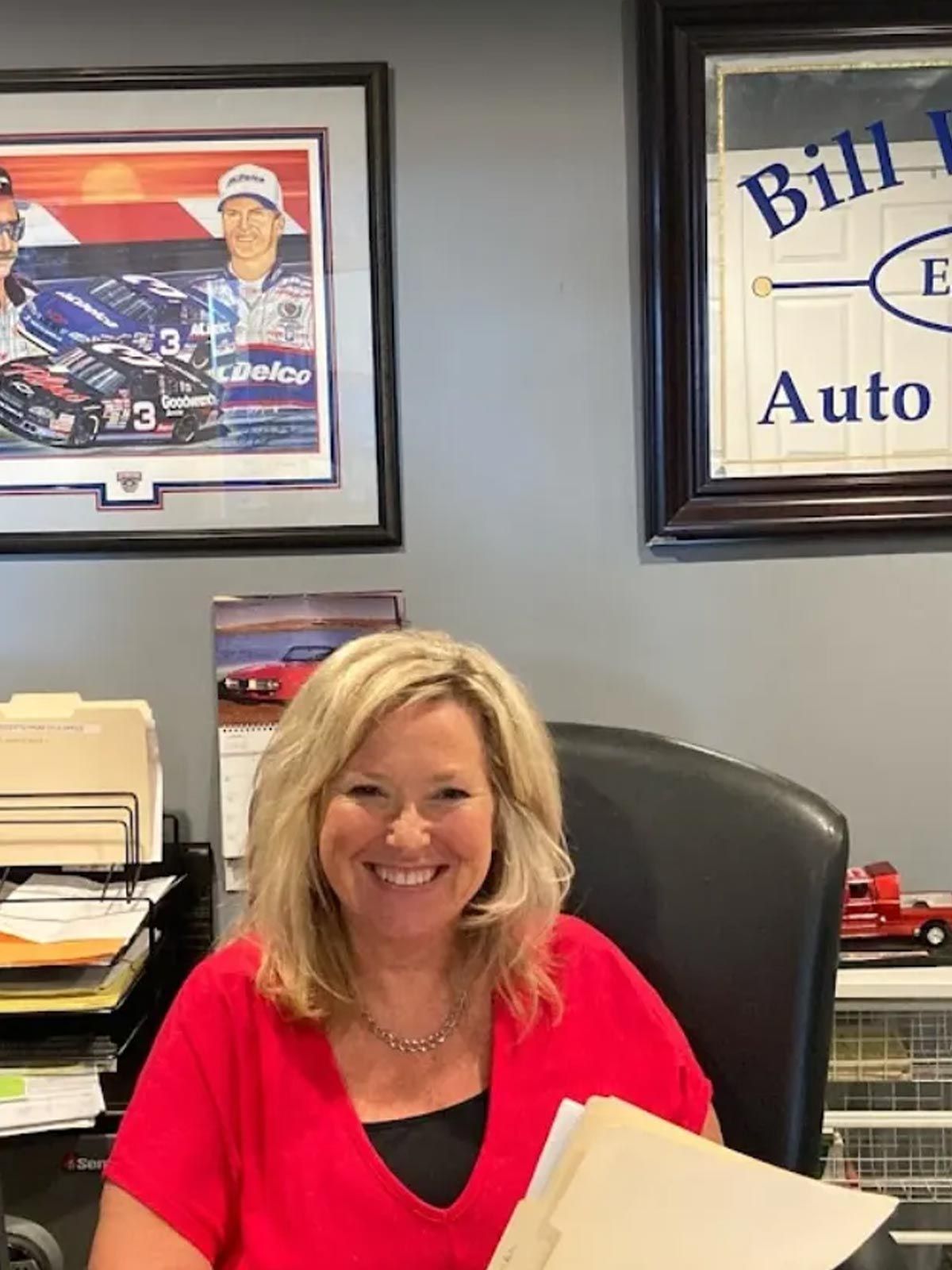 Smiling woman in a red top seated at an office desk with framed posters on the wall behind her