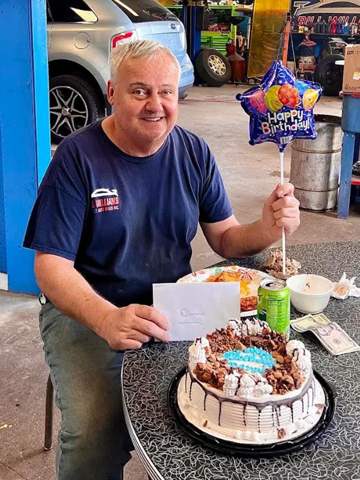 Man smiling at table with birthday cake and blue Happy Birthday balloon, holding a card in a garage.