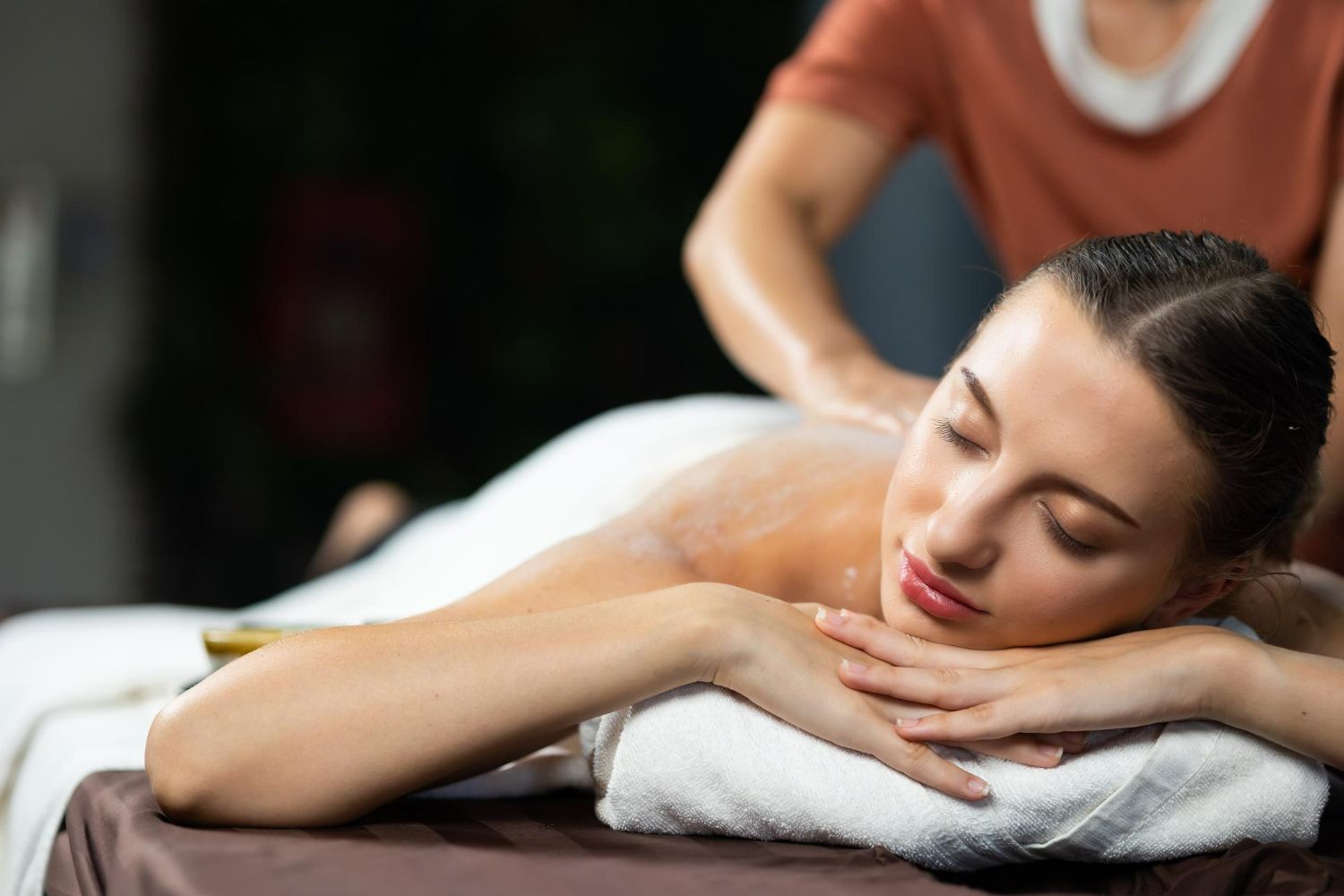 A person lying prone on a massage table with their eyes closed, resting their head on a white towel while receiving massage.