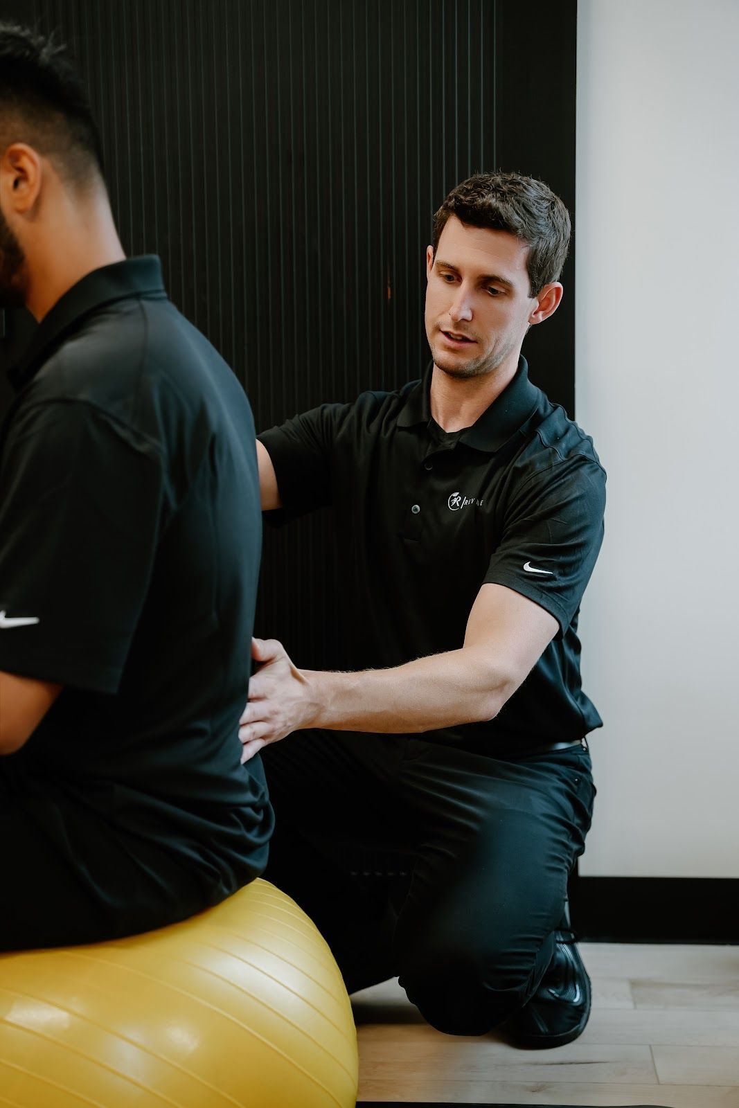 Physical therapist assisting a person seated on a yellow exercise ball, examining their back. Black wall in background.